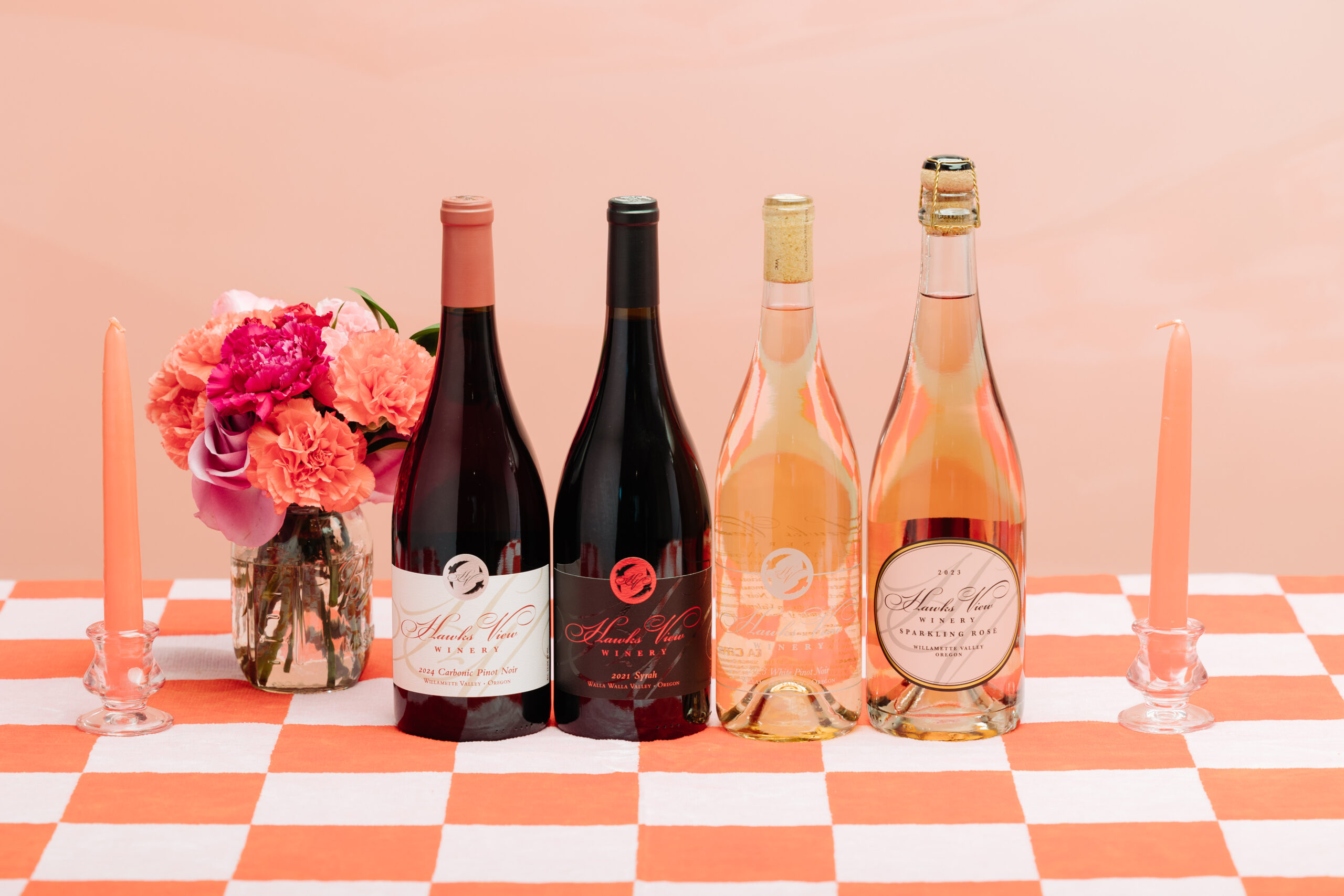 Summer 2024 Allocation bottles. 4 wines bottles from Hawks View Winery sitting on a checkered table cloth between pink flowers and candlesticks.